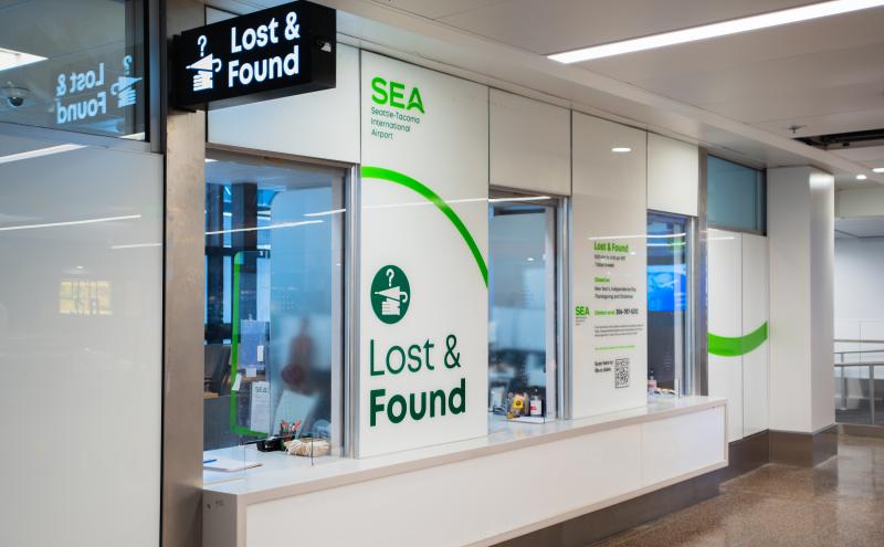 A Lost and Found service area at SEA Airport, with a bright overhead sign reading “Lost & Found” and a large wall display featuring the SEA airport logo and contact information. The space includes service windows, counters with small items, and glass partitions reflecting light from the terminal.