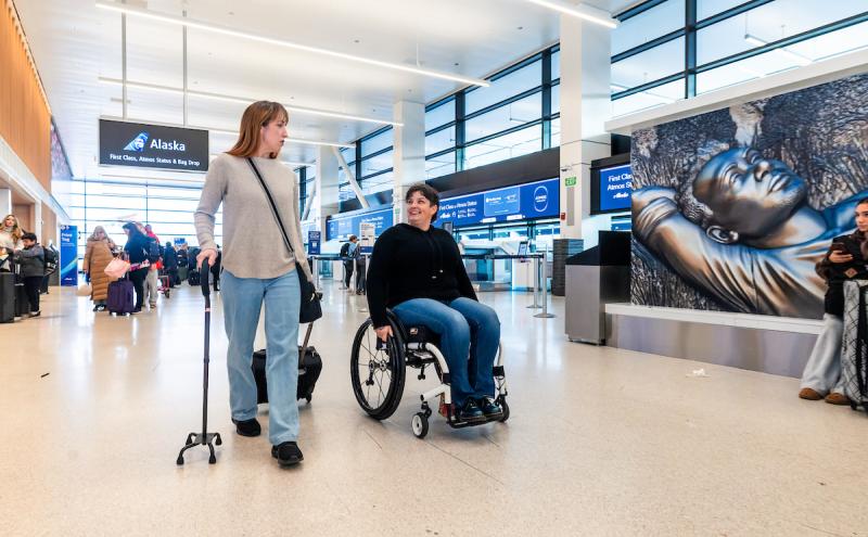 Traveler in a wheelchair and traveler with a cane walking down ticketing