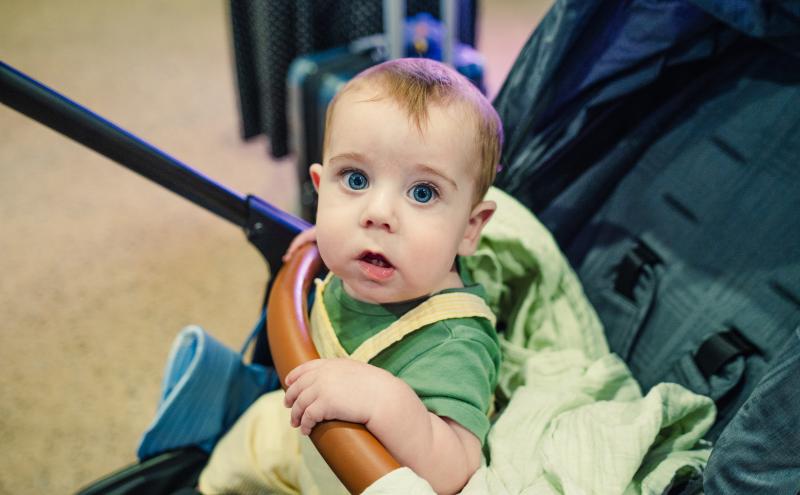 A small child sits in a stroller indoors, holding onto a brown handlebar and covered with a light green blanket. Luggage and other travel items are visible in the background.