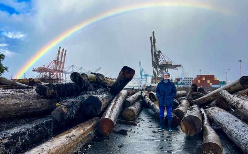 George Blomberg stands at a Port habitat restoration site among large shoreline logs, with cargo cranes and stacked shipping containers in the background. A bright rainbow arcs across the cloudy sky above the terminal.