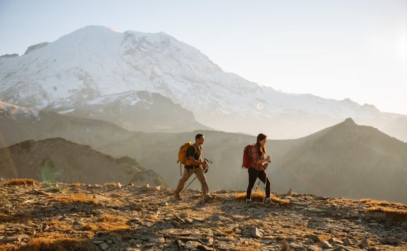 Two hikers during sunset on a trail with a snowy mountain behind them.