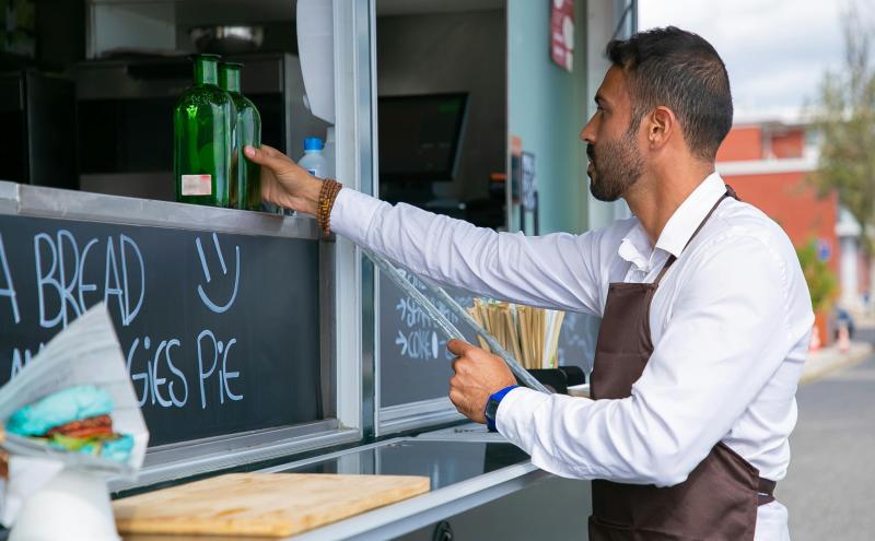 A person wearing a white shirt and apron stands at a food truck counter, reaching for two green glass bottles. A chalkboard menu with handwritten text and a smiley face is visible, along with a wrapped sandwich and a wooden cutting board on the counter. The food truck’s interior and the street outside are in the background.