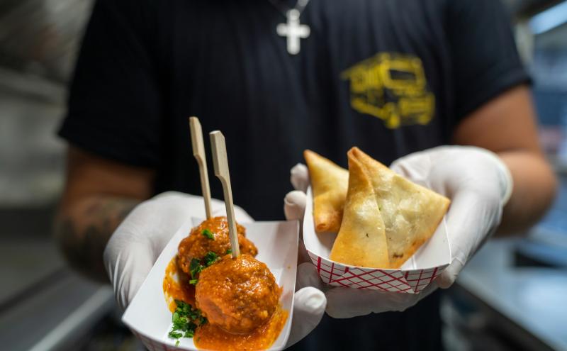 A person wearing gloves holds two small food‑tray servings inside a food truck, one containing sauced meatballs with wooden skewers and the other containing two fried pastry pockets.