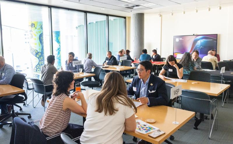 Several groups of people sit at tables in a bright meeting room, engaged in conversations. Signs with organization names, including ‘Casne’ and ‘WSP,’ are placed on some tables. Large windows with patterned columns are in the background, along with a wall‑mounted screen.