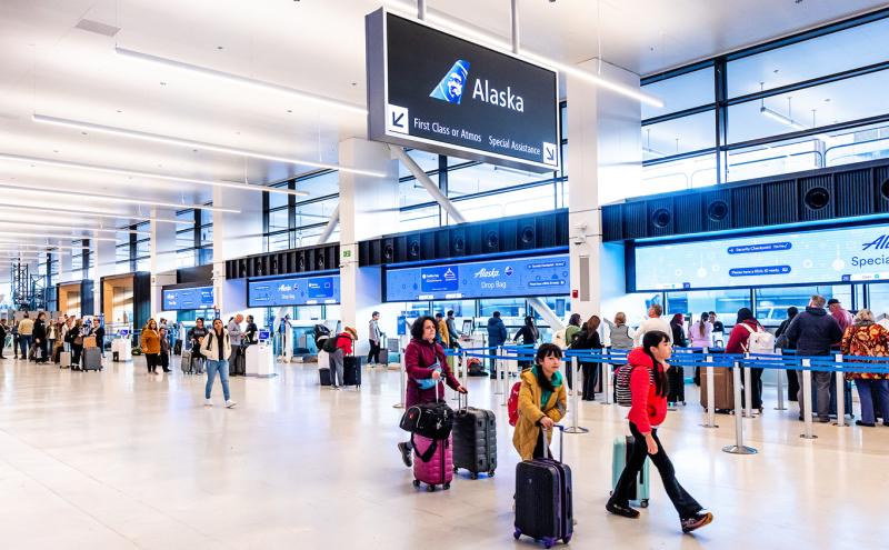 A spacious airport check‑in area with multiple travelers pulling luggage and queuing at self‑service kiosks and counters. Large overhead signs display directions for Alaska Airlines services, including first‑class check‑in and special assistance. Bright lighting and wide walkways are visible throughout the terminal.