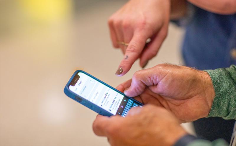 A close-up view of two hands interacting with a smartphone. One hand holds the phone while the other hand points at the screen, which displays a messaging or contacts interface. The background is blurred, placing focus on the hands and the device.