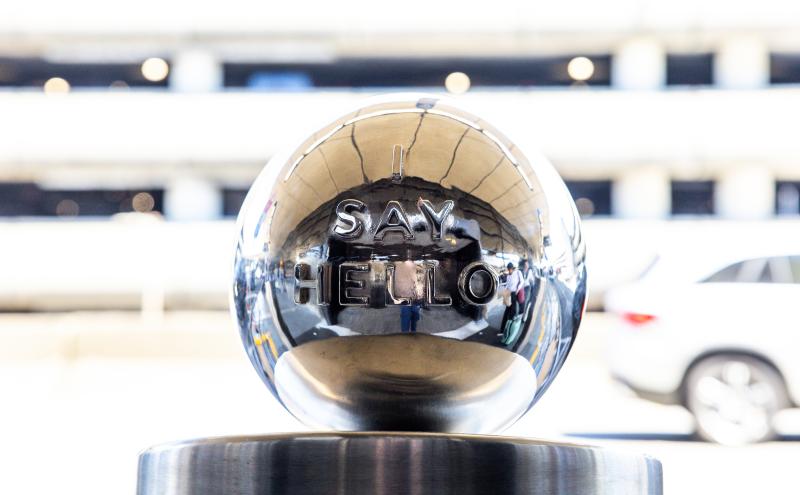 A polished metal sphere with the raised words “SAY HELLO” displayed on its front, reflecting the surrounding outdoor area including cars and people in the background.