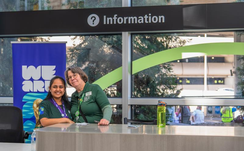 Two staff members stand behind an airport information desk. A sign above them reads ‘Information,’ and a banner beside the desk displays the words ‘We Are.’ A large window behind the desk shows trees and part of the terminal roadway. A green water bottle sits on the counter.