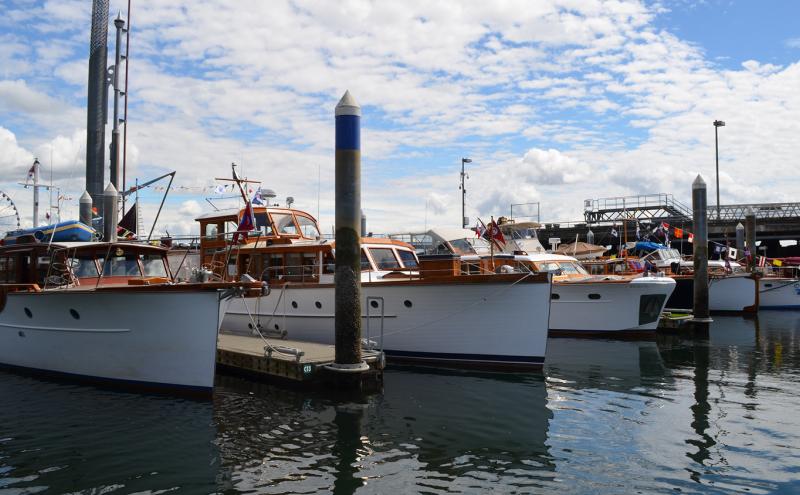 A row of classic wooden motorboats is docked at a marina on calm water, with tall mooring posts in front and a partly cloudy sky above. Colorful signal flags hang from several boats, and a large Ferris wheel and pier structures are visible in the background.