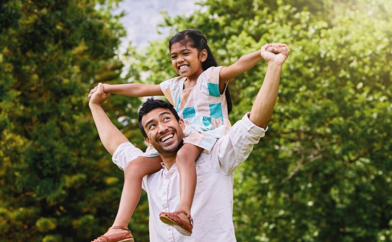 A girl rides on shoulders of her dad as they run through a park
