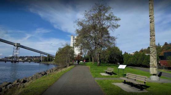 A paved waterfront path runs alongside a rocky shoreline and grassy park area with benches, trees, and a tall carved wooden totem pole. A few people walk in the distance, and an elevated industrial conveyor structure extends over the water under a blue sky with scattered clouds.