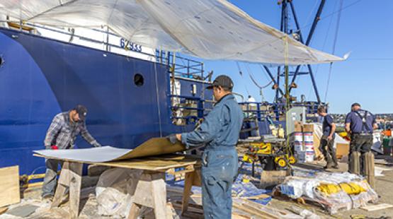 Several people working outdoors on a ship‑side project area, handling large materials and equipment under a temporary plastic canopy.