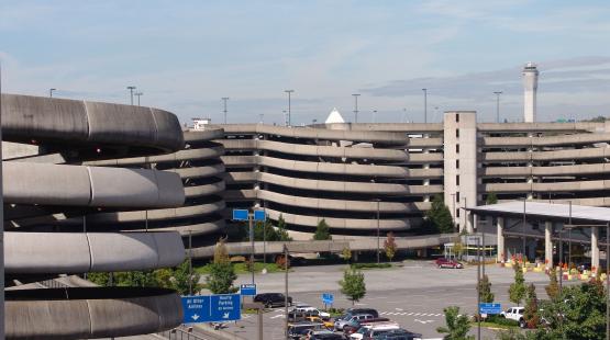 A large concrete parking garage at an airport with multiple levels of curved ramps is shown on a clear day. In the foreground, several levels of the spiral ramps are visible. In the background, additional parking structures, cars in surface parking areas, roadway signs directing drivers to different parking options, and the airport control tower appear. Trees and landscaped areas are scattered throughout the scene.