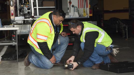 Two workers wearing safety vests and protective gear are kneeling on a concrete floor beside a utility vehicle, examining a small wheeled inspection device placed between them. Tools, equipment, and storage compartments are visible in the background.