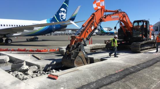 Construction equipment and workers breaking up concrete on an airport tarmac near a parked airplane, with debris and tools visible around the work area.