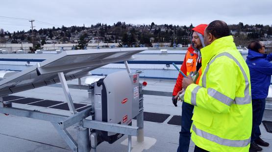 Two workers in safety gear examine rooftop solar equipment at Fishermen’s Terminal, with solar panels and surrounding buildings visible under an overcast sky.
