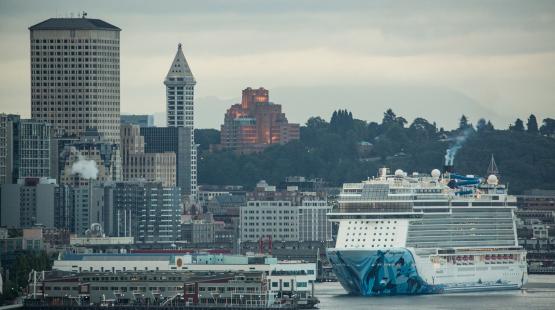 A large cruise ship approaches the Seattle waterfront, with downtown buildings and a cloudy sky in the background.