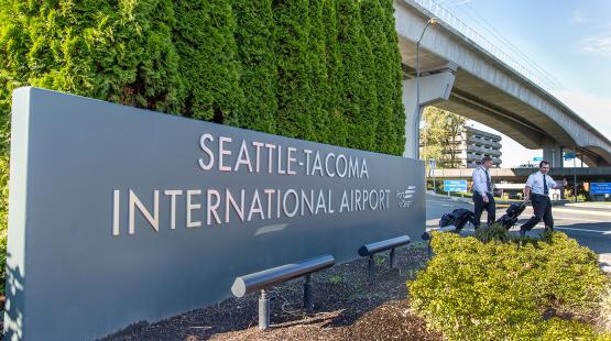 Seattle‑Tacoma International Airport entrance sign with two uniformed airline crew members walking past it, with an elevated roadway and tall greenery in the background.