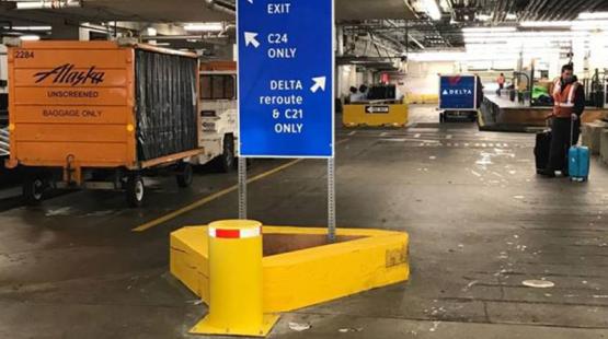 Underground airport service area with a large blue directional sign showing exits and baggage cart traffic routes, surrounded by baggage carts, equipment, and a worker standing near luggage.