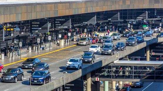Airport terminal roadway with multiple lanes of vehicles picking up and dropping off passengers, and people walking along the curbside outside the terminal building.