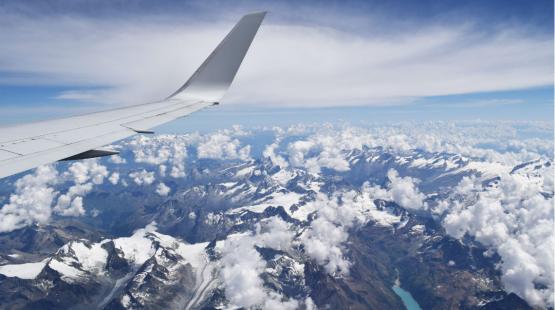 View from an airplane window showing the wing above snowy mountain peaks and scattered clouds.