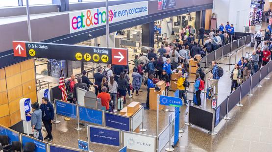 A busy airport security area with long lines of travelers moving through queue lanes near signage for gates, ticketing, and CLEAR kiosks.