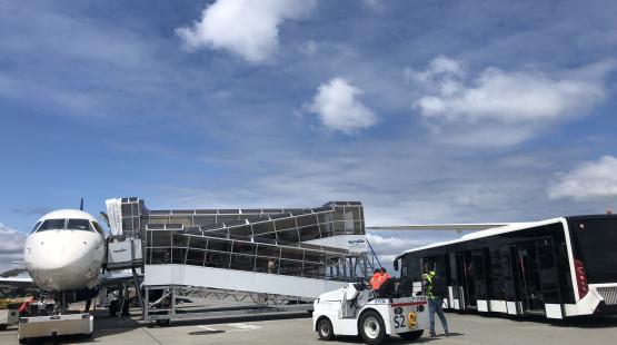 An aircraft parked on the airport ramp with a mobile boarding ramp attached, an airport bus positioned nearby, and ground crew working beside a small tow vehicle under a partly cloudy sky