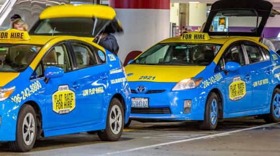 Two bright blue-and-yellow taxis labeled ‘For Hire’ are parked at a curb, with one vehicle’s trunk open as someone loads or unloads items.
