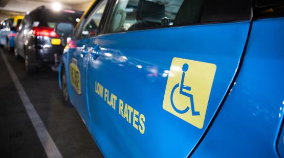 A row of taxis parked in a covered area, with a bright blue taxi in the foreground displaying a wheelchair accessibility symbol and the words “LOW FLAT RATES” on its side.