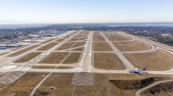 Aerial view of multiple Sea Airport runways with a commercial airplane taxiing and airport buildings and landscape visible in the background.