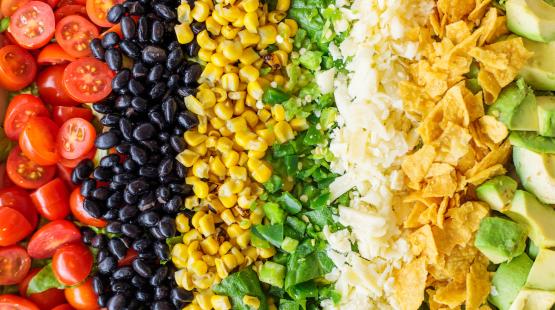 A close-up view of neatly arranged salad ingredients in horizontal rows, including sliced cherry tomatoes, black beans, corn kernels, chopped green bell peppers, shredded cheese, crushed tortilla chips, and diced avocado.