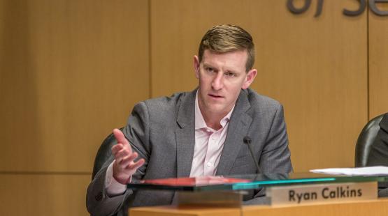 Commissioner Calkins sits at a meeting table during a Commission meeting, gesturing with one hand while speaking into a microphone, with a nameplate labeled “Ryan Calkins” visible in front.