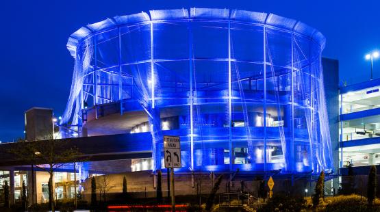 A circular multi-story structure wrapped in hanging strands of wire mesh draped from the roof to the ground, lit from within at night. 