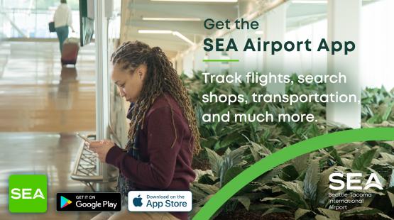 A person with long braided hair stands in an airport terminal holding a smartphone near a seating area. Large green plants line the right side of the space, and another traveler with luggage is visible in the background. Text on the image promotes the SEA Airport App, stating that it can track flights, search shops, find transportation, and more. Logos for SEA, Google Play, and the App Store appear at the bottom.
