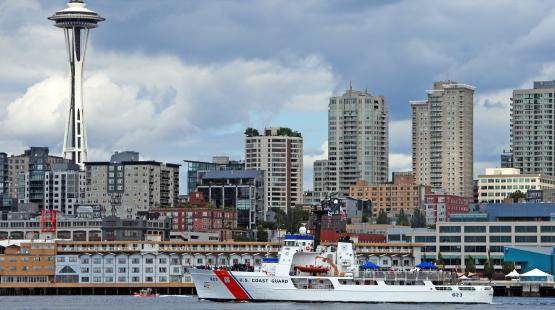 A large U.S. Coast Guard vessel travels across a body of water with a city skyline in the background. The skyline includes several tall modern buildings and a distinctive tower with a wide circular top. The sky is cloudy above the waterfront scene.