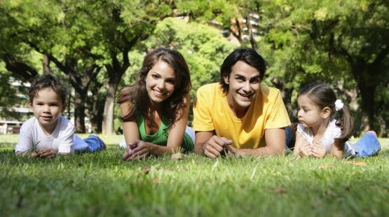 Four people lie on the grass in a park surrounded by tall trees. Two adults are positioned in the center, with one child on each side. All four individuals are facing the camera while resting on their elbows, with sunlight filtering through the trees in the background.