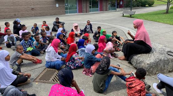 A group of children seated outdoors in a semicircle on a paved area, listening to an adult who is sitting on a large rock and speaking to them. A building with windows and doors is in the background, along with a bench and grassy area.