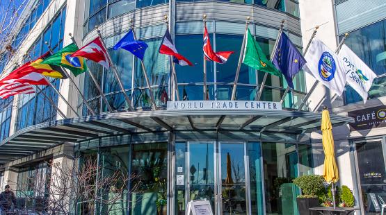 Exterior view of the World Trade Center Seattle building, featuring a curved glass façade and a row of international and organizational flags displayed above the main entrance. The entrance canopy, reflective windows, and nearby café umbrellas and signage are visible.