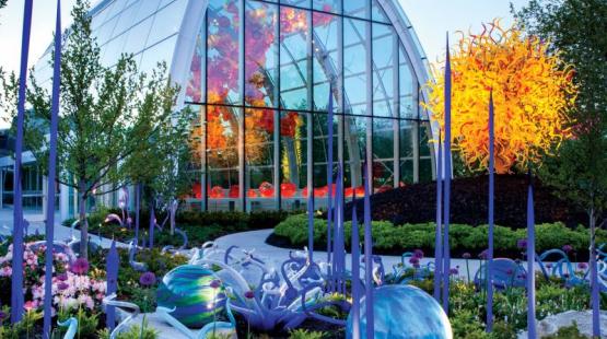 Outdoor garden featuring colorful glass sculptures among flowers and greenery. In the background, a large glass conservatory displays vibrant orange and red glass artwork inside, reflected on its curved windows. In the foreground, tall slender purple glass forms and rounded blue‑green glass shapes are arranged within landscaped plant beds.