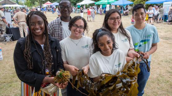 A group of people stands outdoors at a community event, holding large pieces of seaweed. Behind them are booths, tents, and other attendees in an open grassy area.