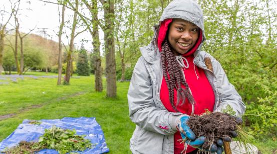 A community member wearing outdoor work clothes holds a bundle of plants and soil while working in a green, wooded area during a Duwamish habitat restoration activity.