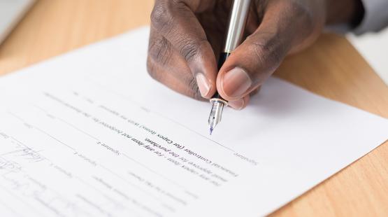 Hand signing a document on a light wood desk.