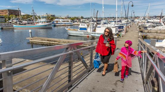Two people walking up a metal ramp from a marina, carrying bags, with docked fishing boats and sailboats in the background under a bright, partly cloudy sky