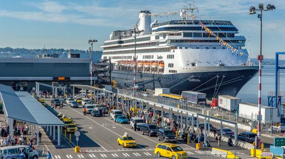 Large cruise ship docked at a busy terminal with long lines of taxis, cars, and passengers moving through loading and drop‑off areas under covered walkways.