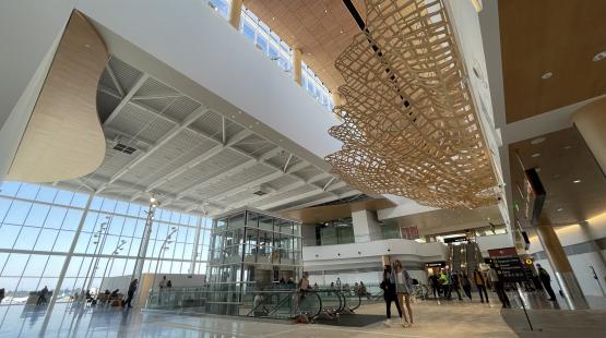 Spacious airport terminal with large windows, escalators, and a hanging wooden sculpture.