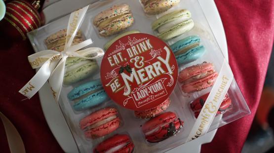 Box of assorted colorful macarons with a round red label reading ‘Eat, Drink & Be Merry,’ tied with a cream ribbon and displayed among holiday decorations.