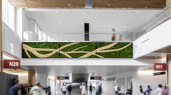 Airport concourse with travelers walking below a large green wall art installation featuring tan, branch‑like patterns. Overhead is a glass‑railed walkway, and signs for gates N20 and N11 are visible.