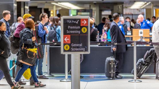 Airport security area with travelers lining up near a sign directing passengers to TSA PreCheck premium screening or general screening.