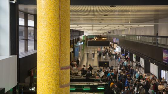 A mosaic-covered column rising the full height of the airport concourse, with a busy terminal visible behind it.