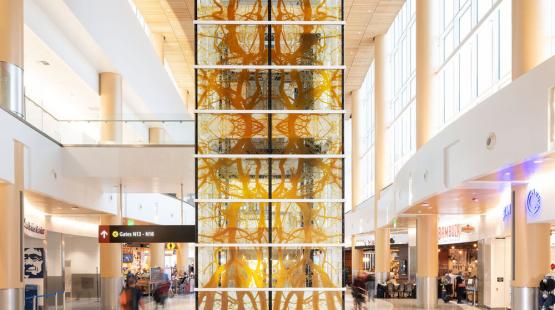 A tall narrow column of glass panels rising the full height of the airport consourse, etched with branching root and limb forms spreading across the surface.